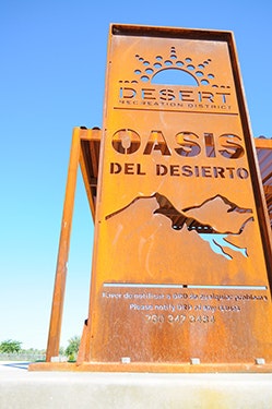 A metal sign for "Oasis Del Desierto" in the Desert Recreation District, featuring a sun and mountain design.