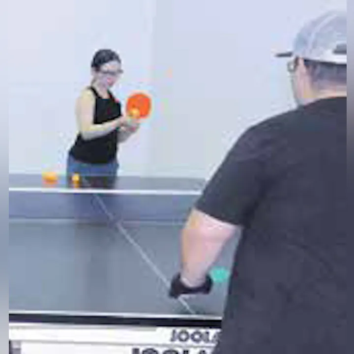 A person with glasses plays table tennis, preparing to hit a ball while another person watches or prepares to respond.