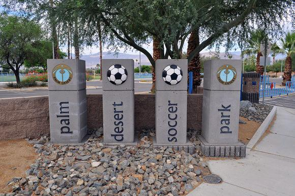 The image features stone pillars marking "Palm Desert Soccer Park," decorated with soccer balls and local emblems.
