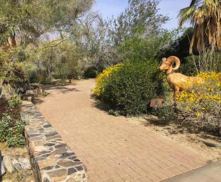 A winding pathway through lush greenery and colorful flowers, featuring a decorative ram sculpture on the side.