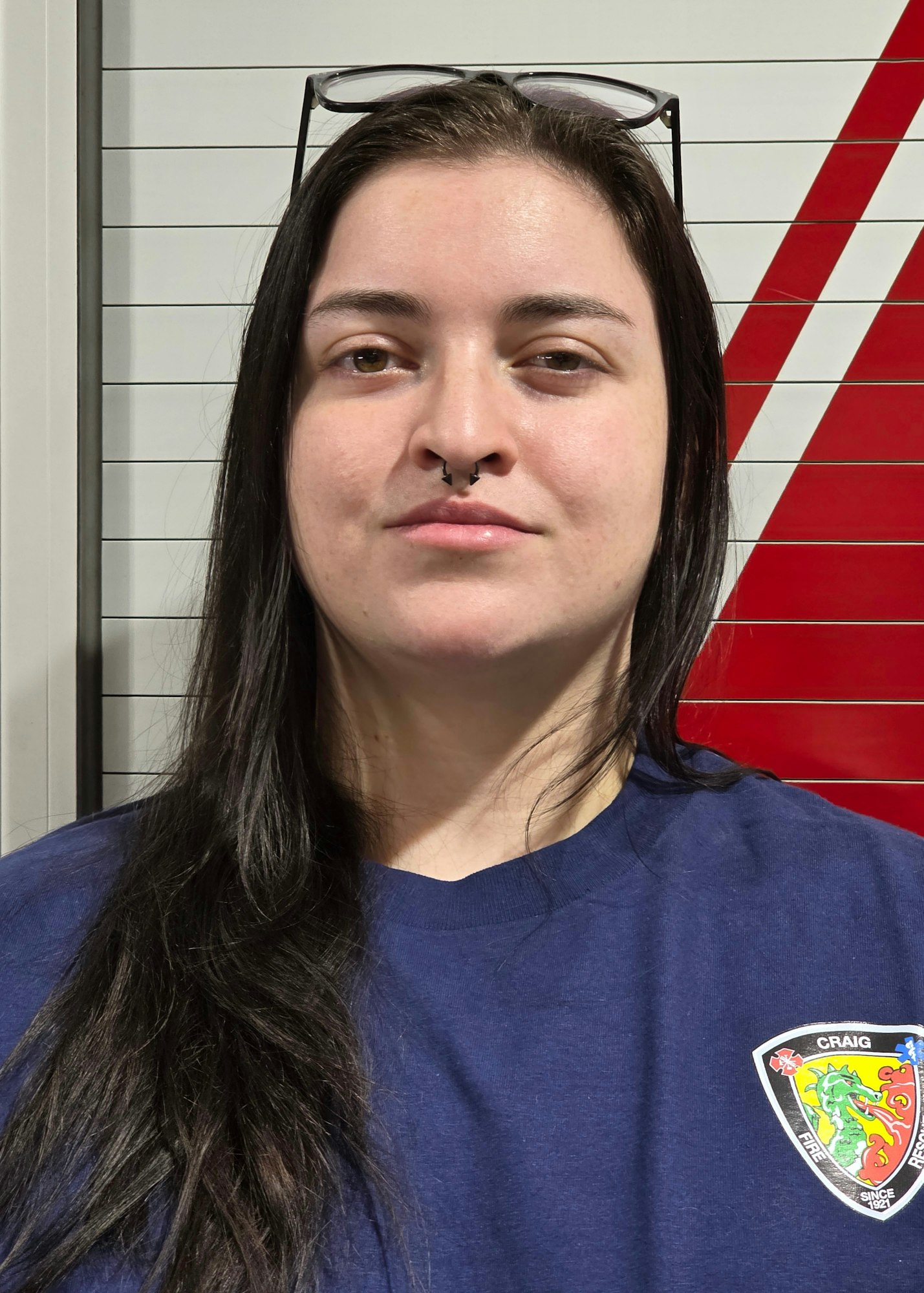 A person with long dark hair, a septum piercing, and a navy shirt with a logo stands in front of a textured background.