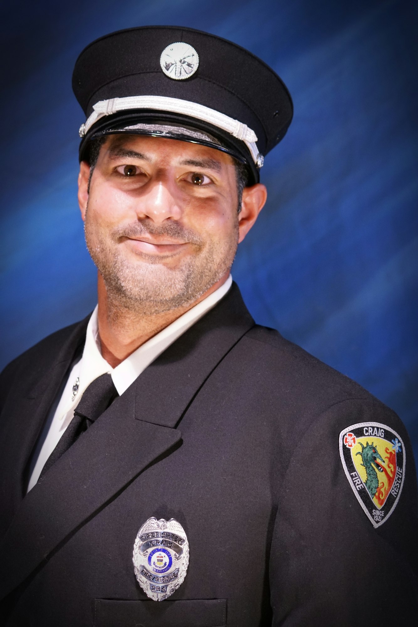 A smiling person in a formal uniform with a badge and a patch, suggesting they may be a service member, such as a firefighter.