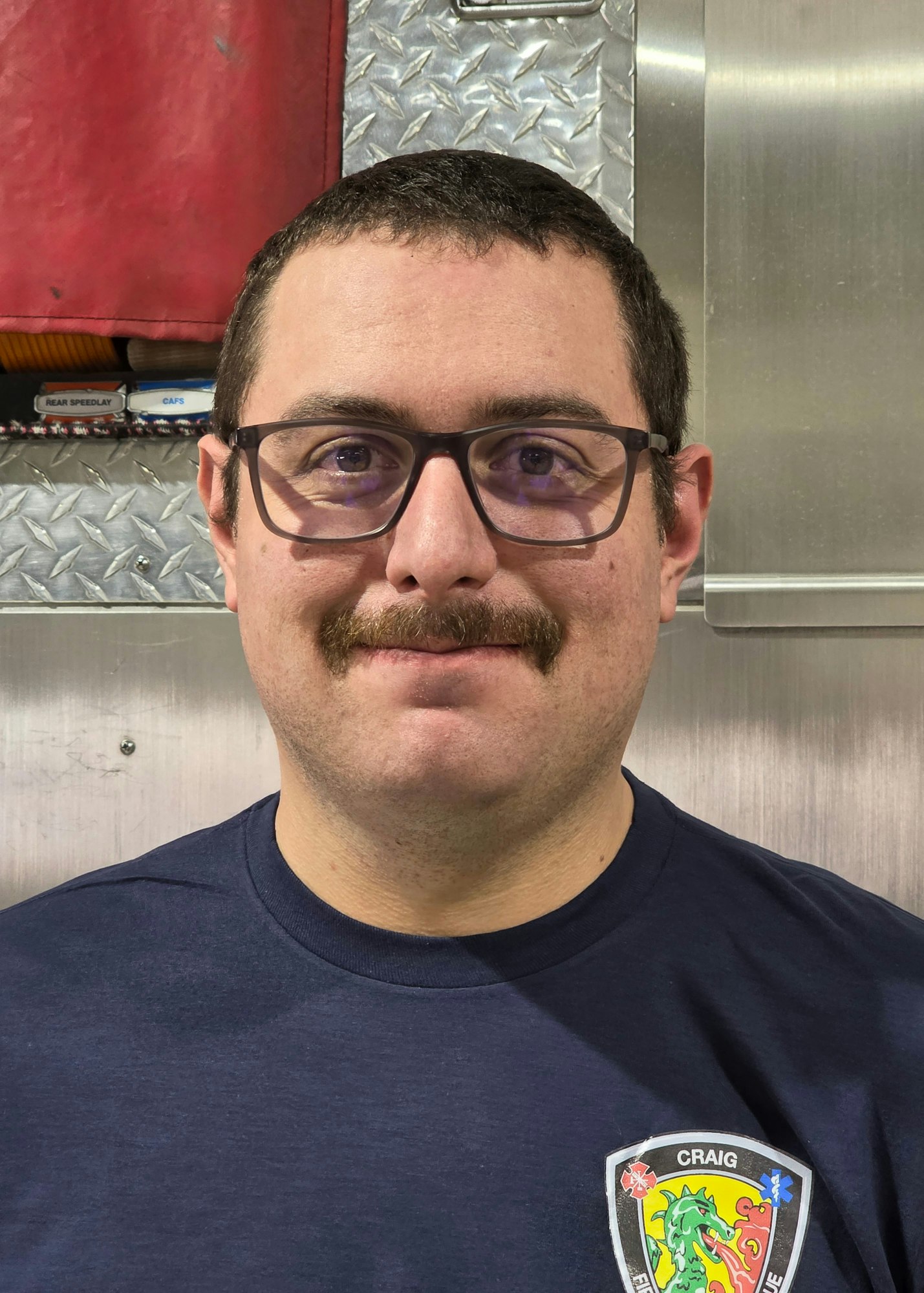The image features a man with glasses and a mustache, wearing a navy shirt with a patch, in a metallic setting.