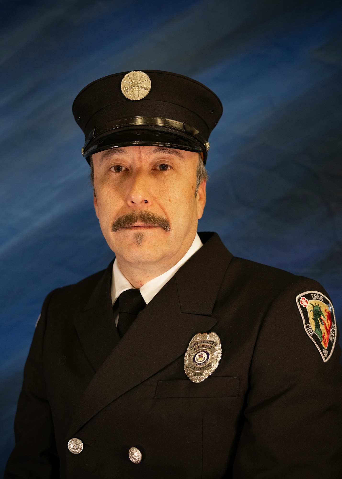 A man in a firefighter's uniform and cap poses for a portrait against a blue background. He has a mustache and a badge.