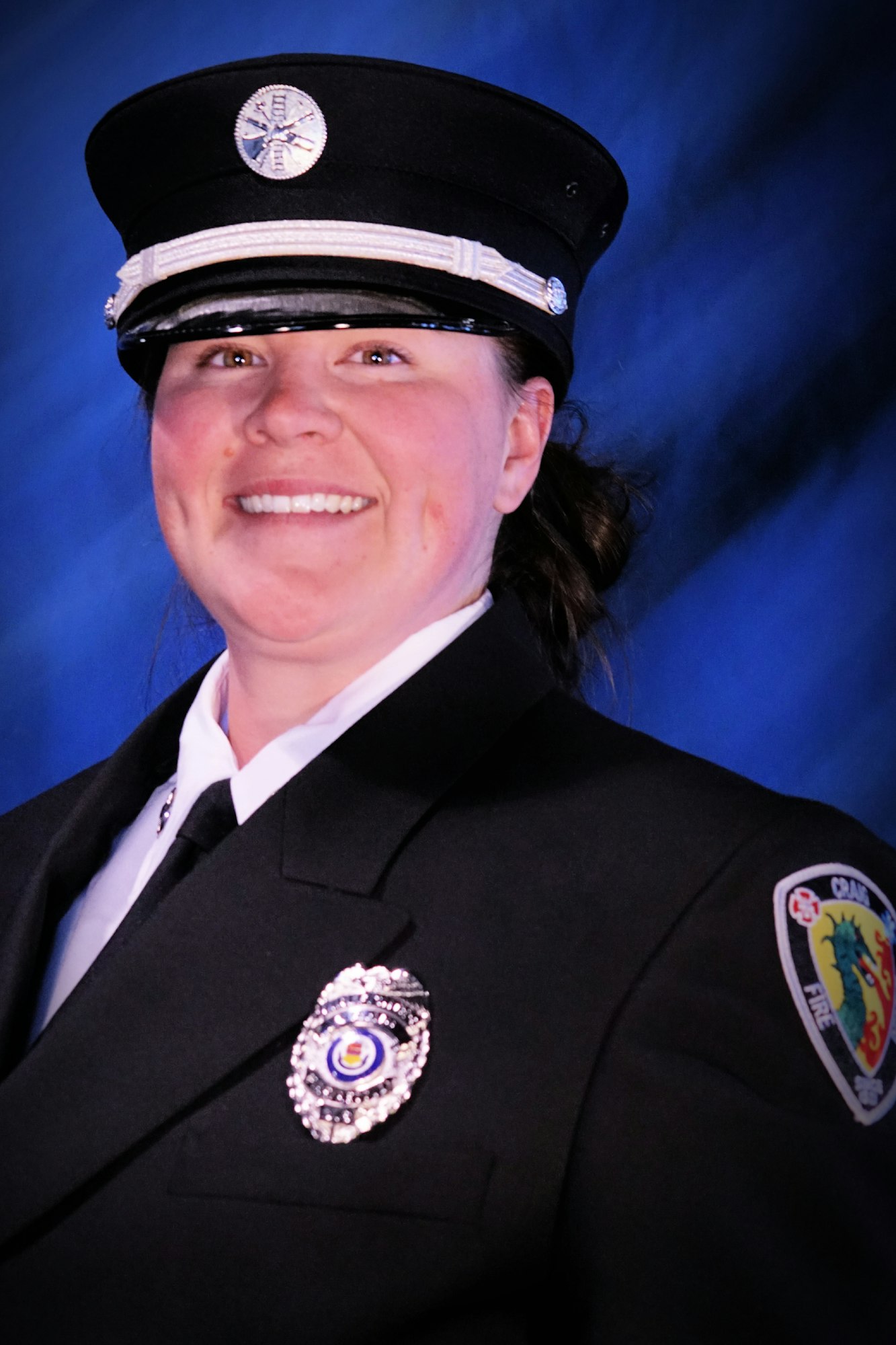 A smiling person in a fire department uniform with badges and a cap.
