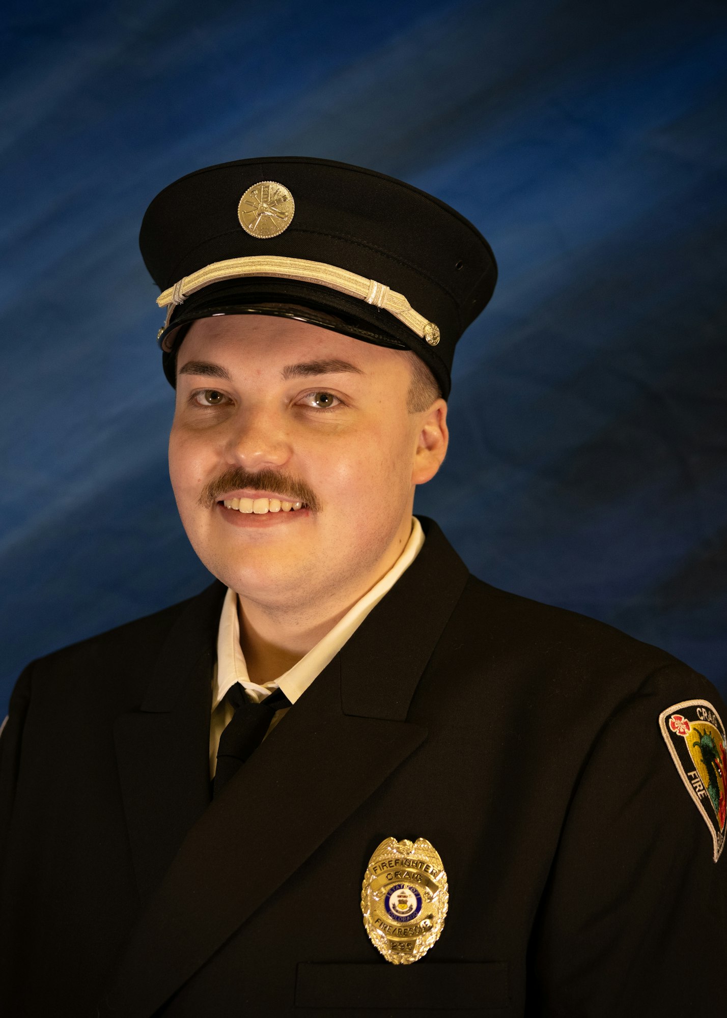 A smiling individual in a firefighter uniform and hat, with a badge, poses against a blue background.