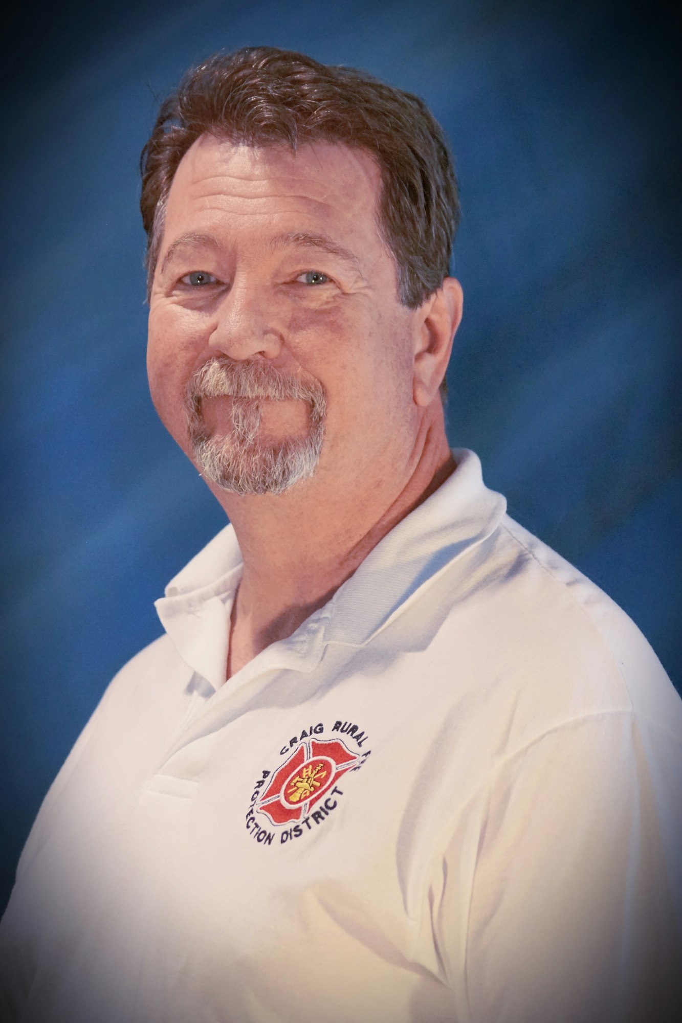 Portrait of a smiling man with a mustache, wearing a white polo with a fire service emblem.