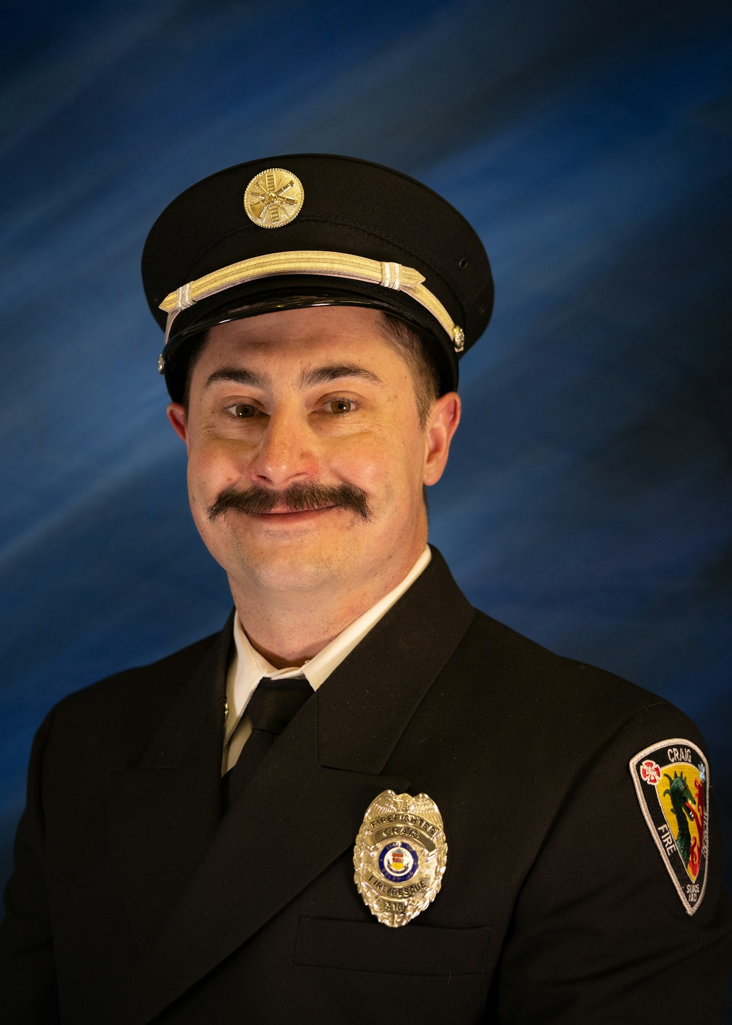 A man in a fire officer uniform smiles for the camera, wearing a badge and hat against a blue backdrop.