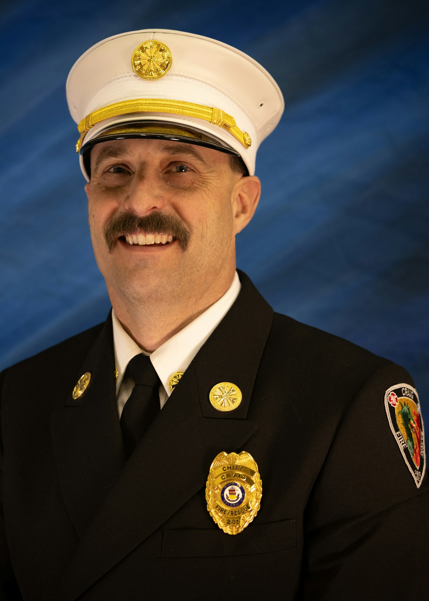 A smiling individual in a formal fire chief uniform, complete with a hat and badge, is posing against a blue background.