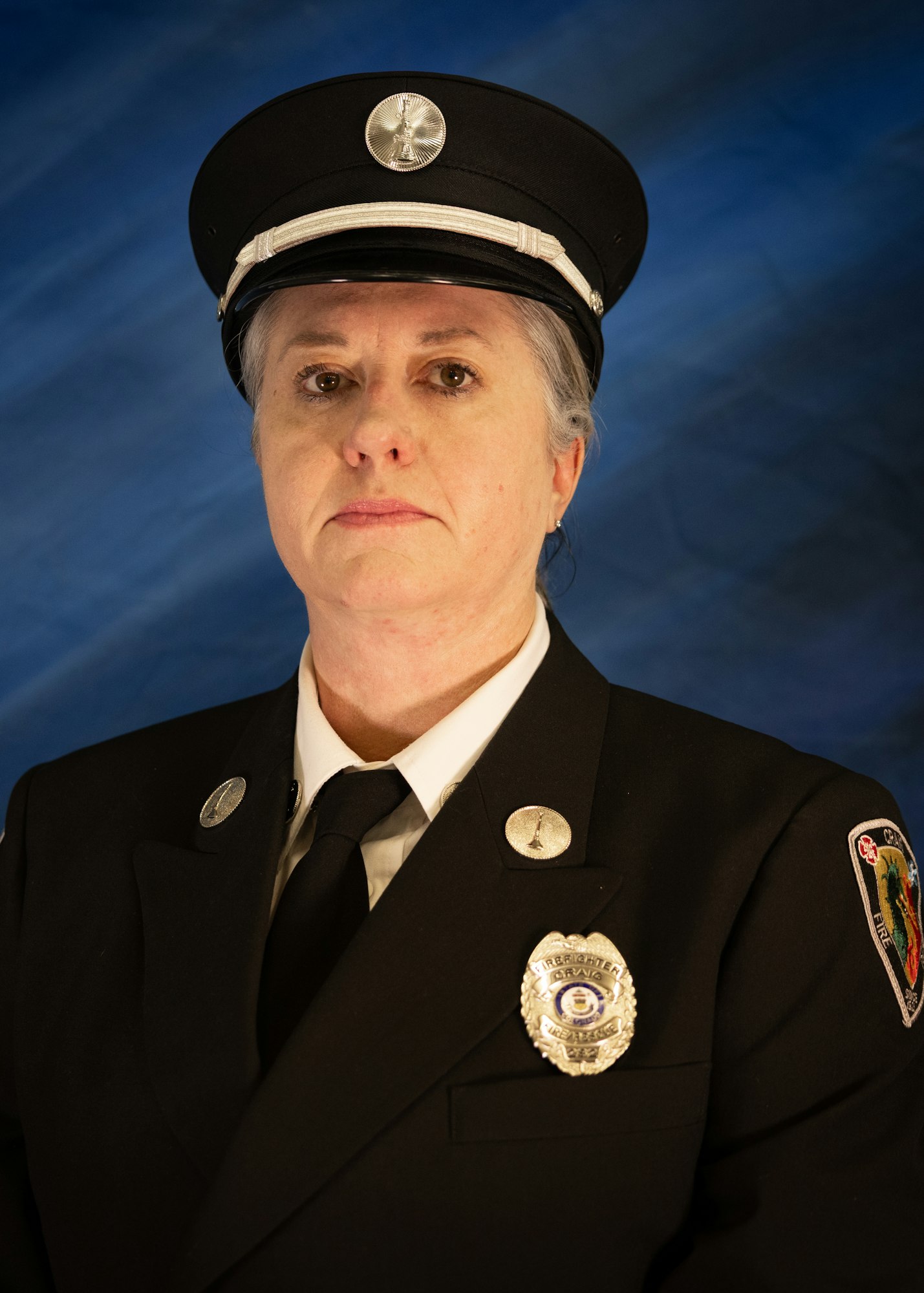 A person wearing a fire department uniform, complete with a badge and hat, poses against a blue background.