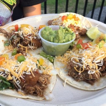 Four tacos topped with shredded cheese and diced peppers, served with a small cup of guacamole on a plate.