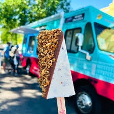 Chocolate-covered ice cream bar with nuts, with a blue and red food truck and people in the background.