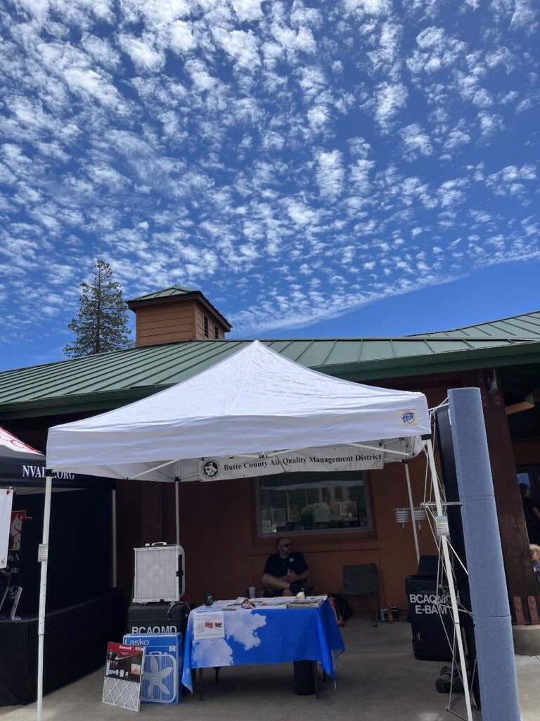 An outdoor booth with banners from "Butte County Air Quality Management District," a clear sky with scattered clouds, and a person seated.