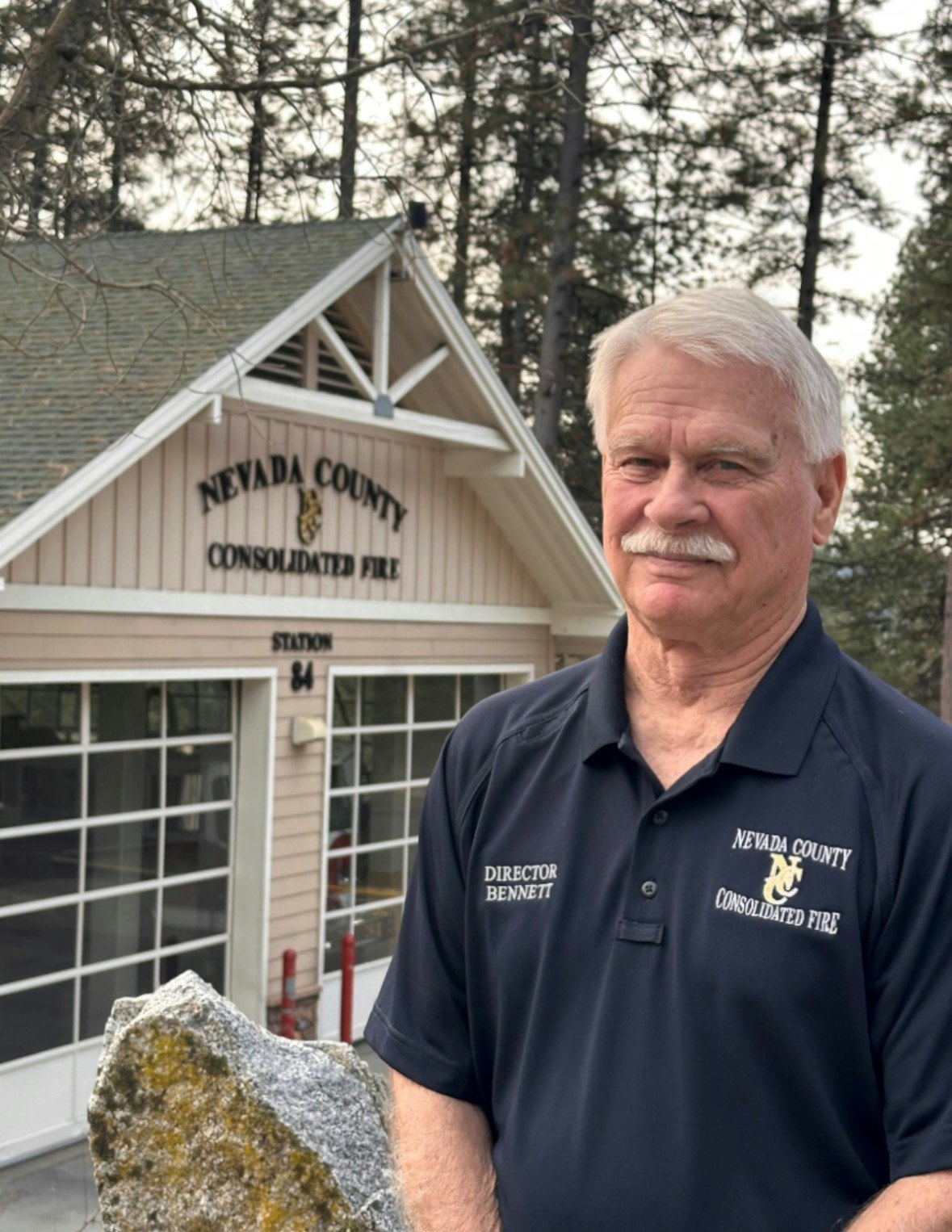 A man in a "Nevada County Consolidated Fire" shirt stands outside a fire station surrounded by trees.