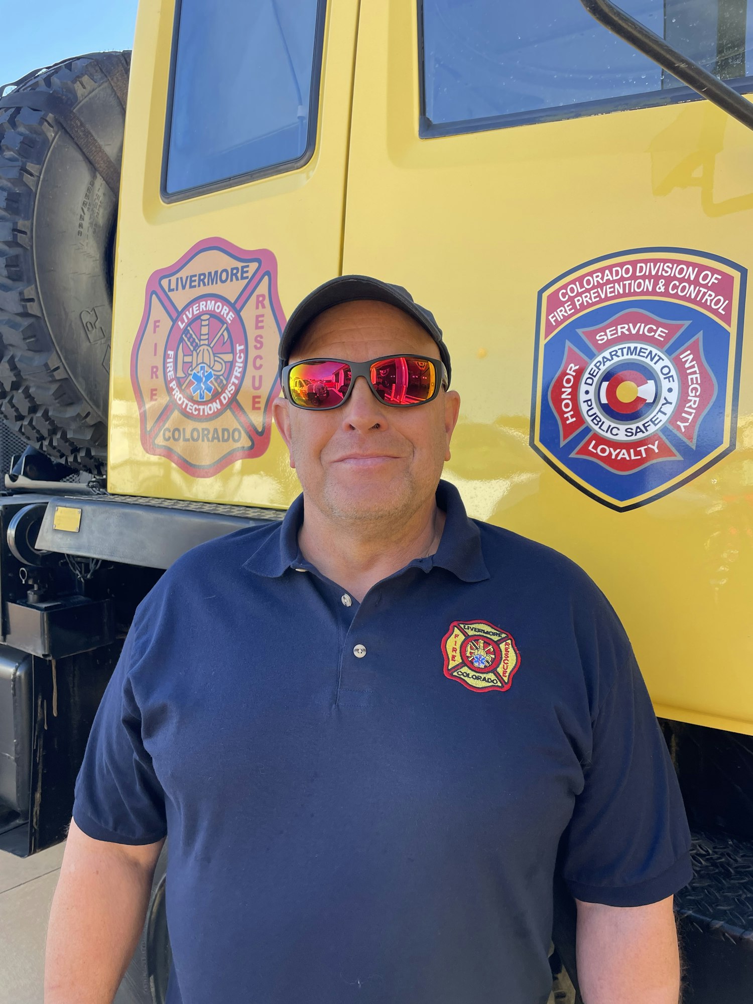 A person in sunglasses is standing next to a yellow vehicle with fire department logos.