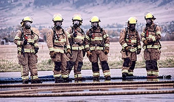 A group of six firefighters in full gear stands together, ready for action, with hoses laid out in front of them.