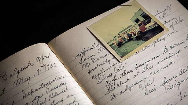 An open notebook from 1938 with handwritten notes and a photo showing cars parked outside a building.