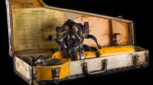 The image shows a vintage gas mask housed in a worn case with instructions and additional equipment.