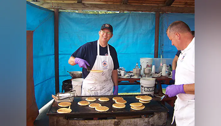 Two men are cooking pancakes on a griddle in a blue-tented area, wearing aprons and gloves, smiling while working together.