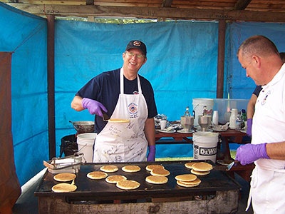 Two men are cooking pancakes on a griddle in a blue-tented area, wearing aprons and gloves, smiling while working together.