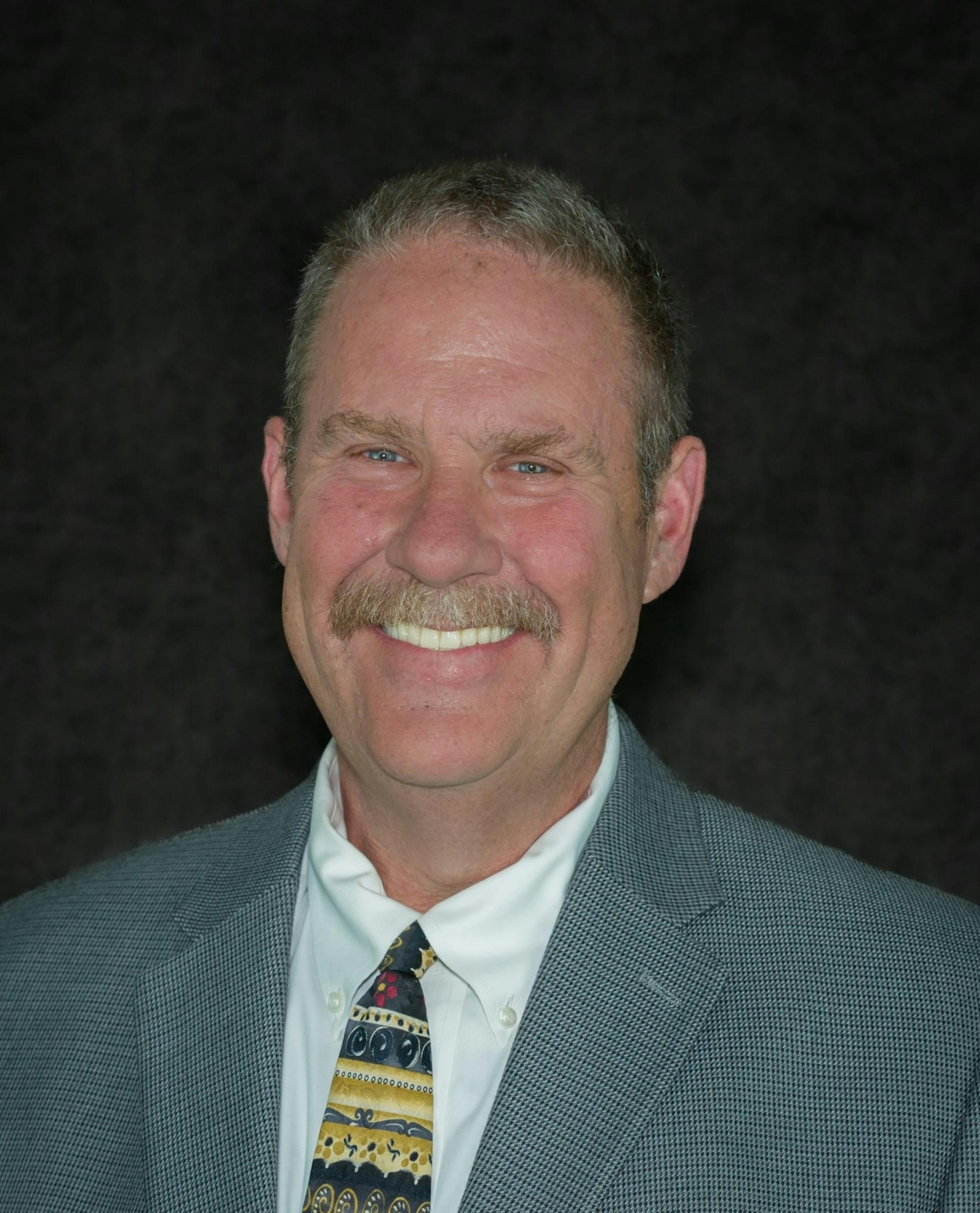 A smiling man with a mustache, wearing a suit and a colorful tie, against a dark background.