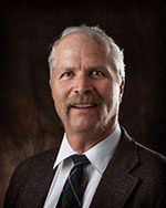 The image depicts a smiling man with a mustache, wearing a suit and tie, against a dark background.