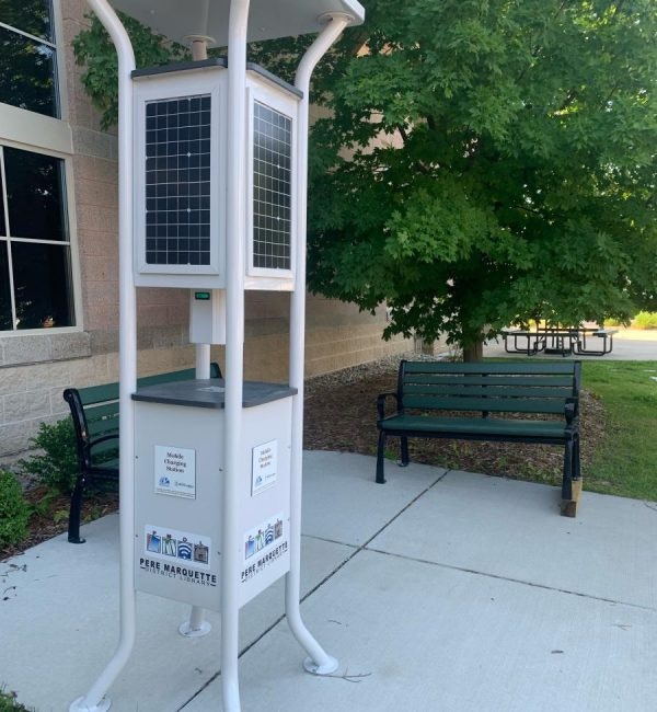 Solar-powered charging station outside a building, surrounded by benches, trees, and a concrete path.