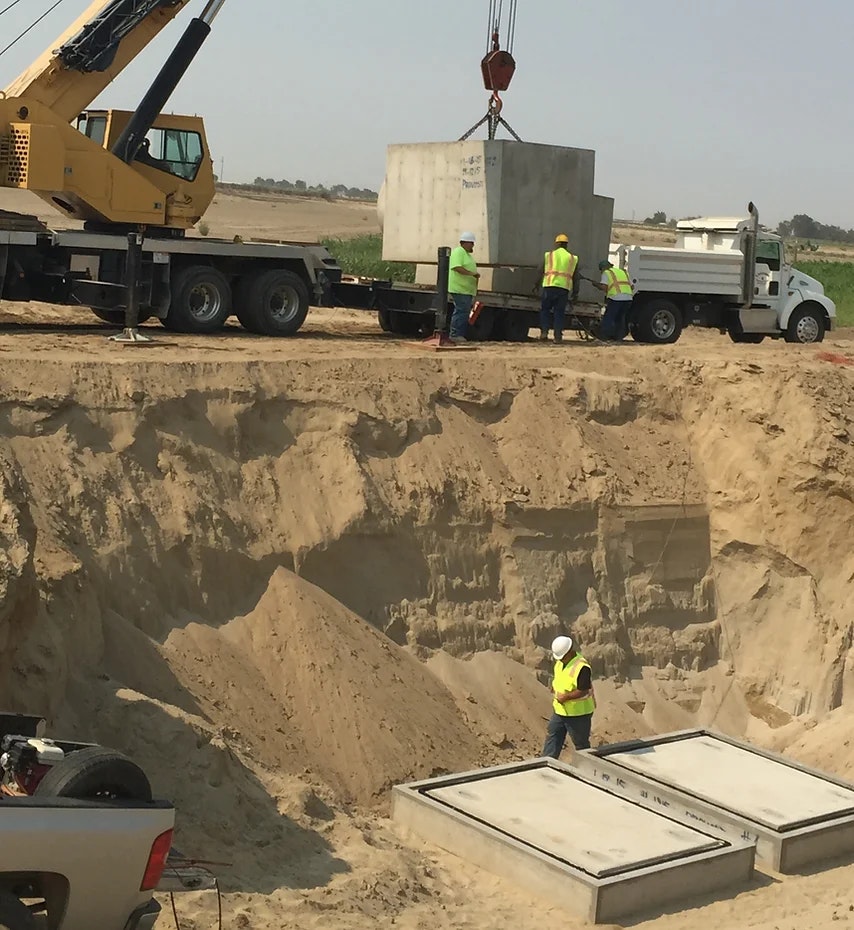 Workers with crane lowering concrete box into excavation site.