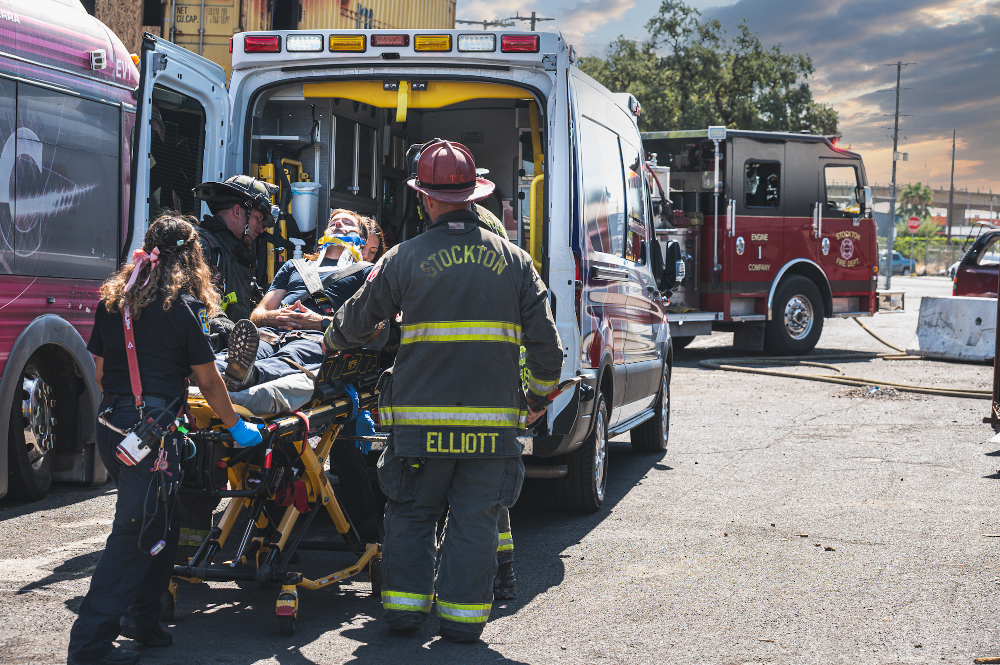 Emergency personnel assist an injured person on a stretcher near a fire truck and ambulance.
