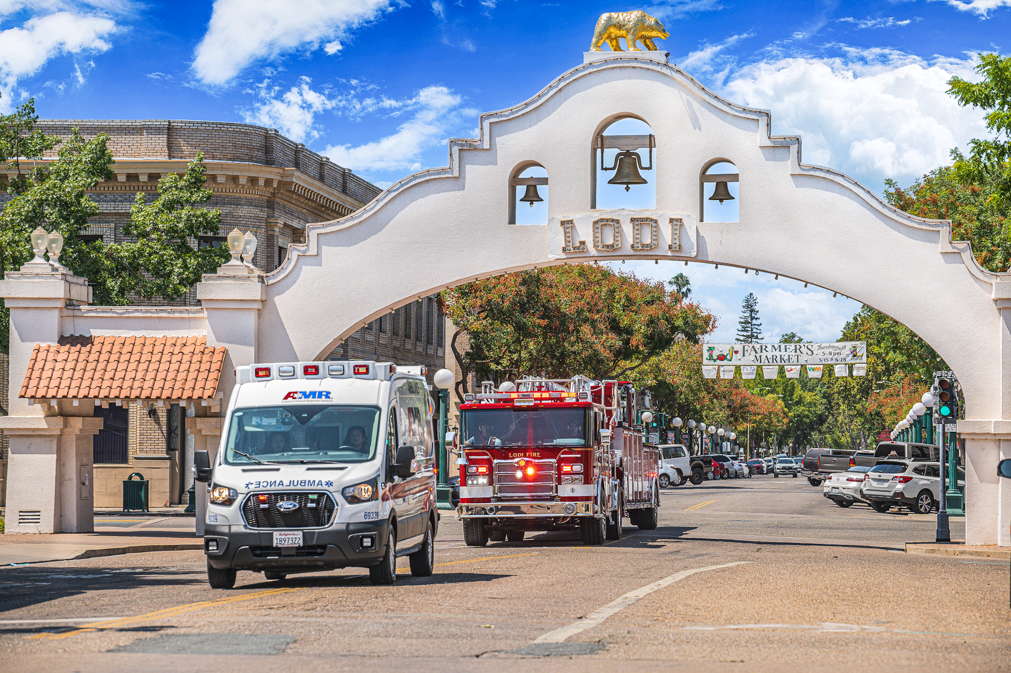 Ambulance and fire truck under Lodi archway, with "Farmers Market" sign and parked cars in the background.