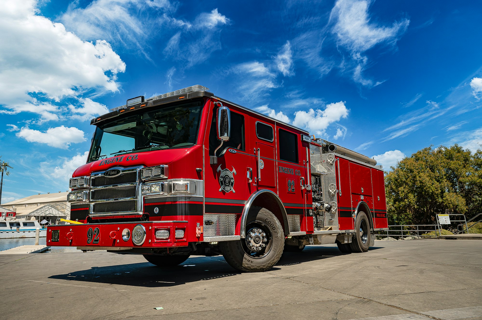 A red fire truck on a sunny day, parked on a road next to trees and under a blue sky with clouds.