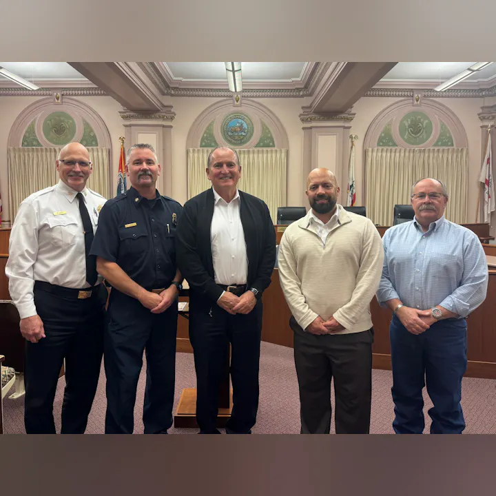 Five men, including firefighters and officials, pose for a photo in a formal setting, likely a government building.