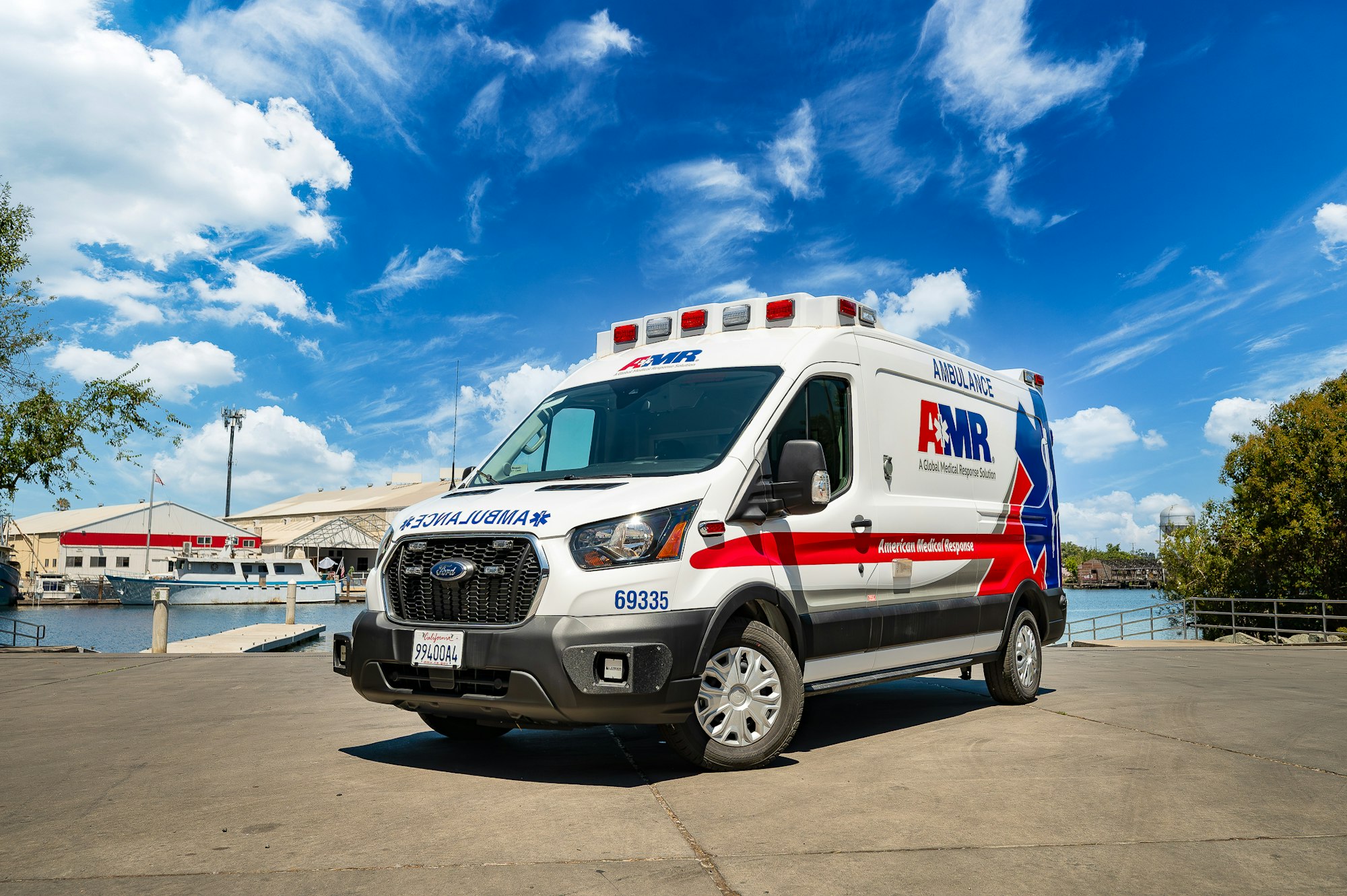 An ambulance with "AMR" branding parked outdoors near a waterfront, under a blue sky with clouds.