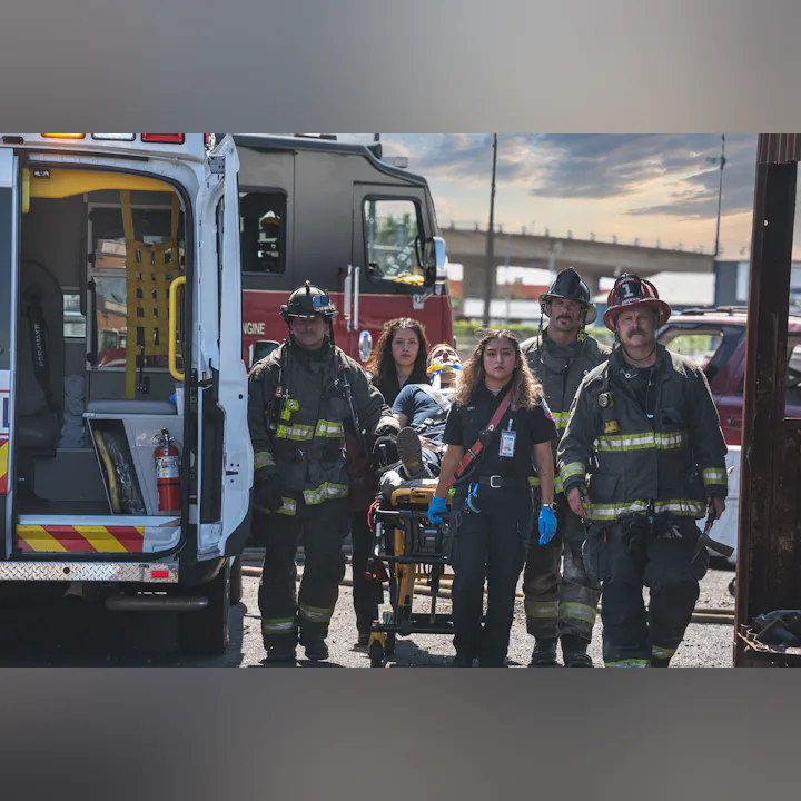 Firefighters and paramedics transporting a person on a stretcher. An ambulance and fire truck are visible.