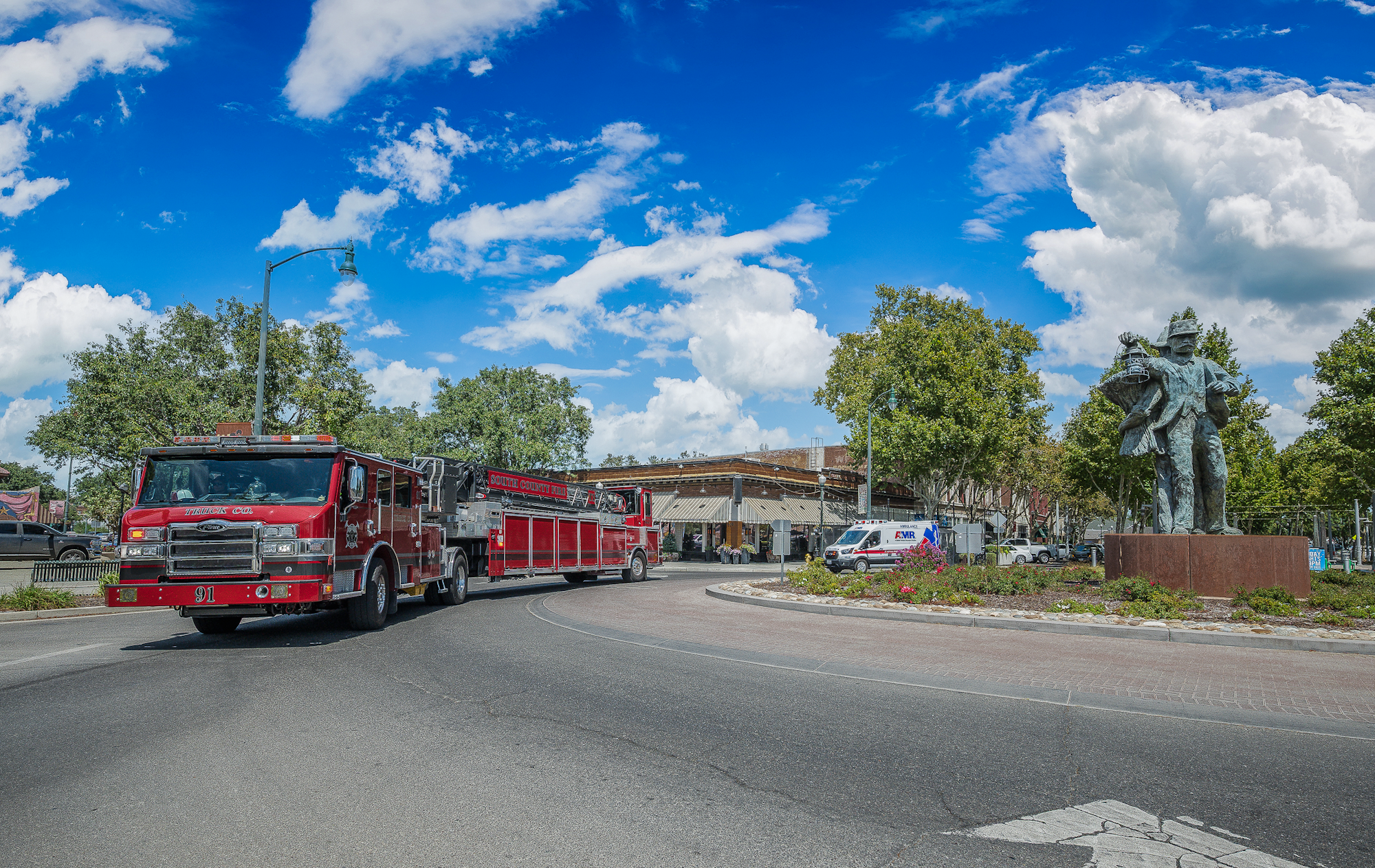 Fire truck and ambulance on a road near a sculpture and green trees under a bright blue sky with clouds.
