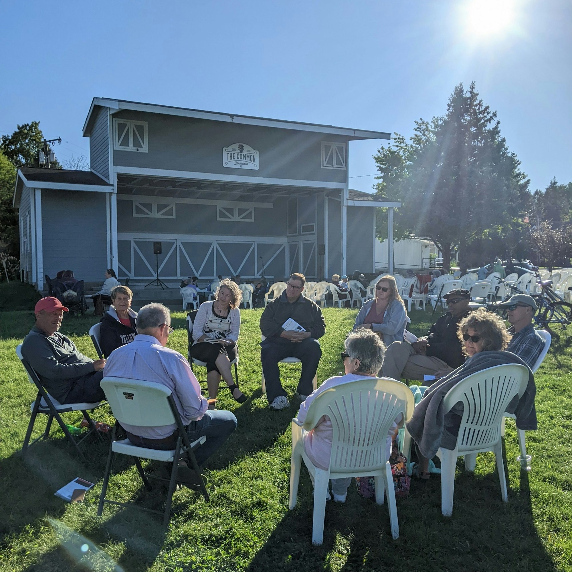 A group of people sit in chairs outdoors, engaging in conversation near a stage in a park setting on a sunny day.