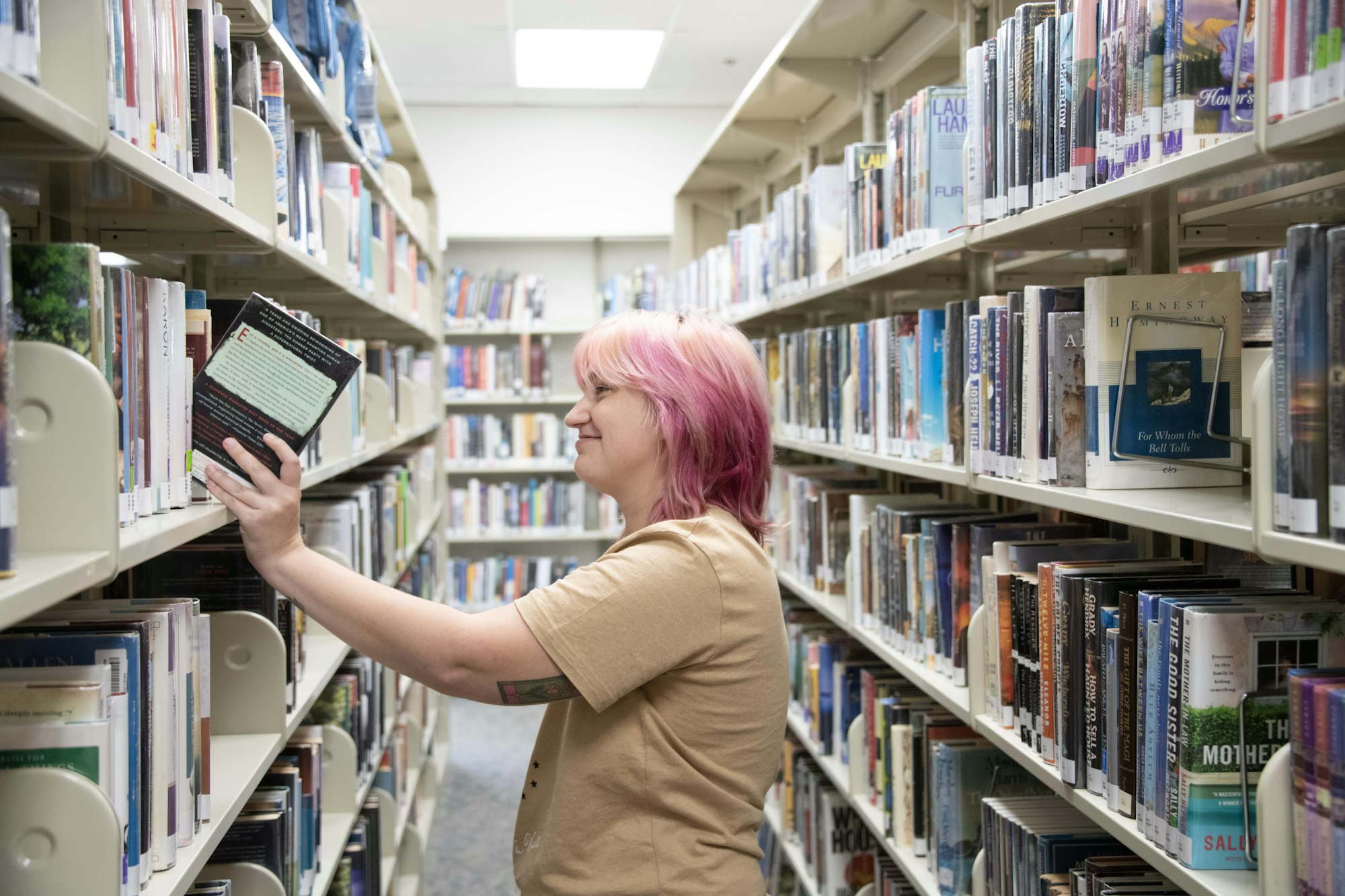 A person with pink hair selects a book from shelves in a library aisle.