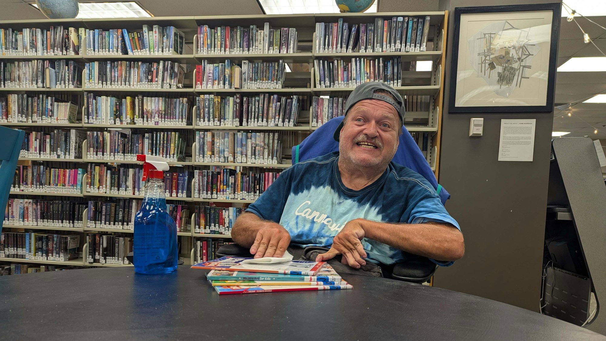 A person smiling, wearing a cap backward, in a library with a spray bottle and books on a table.