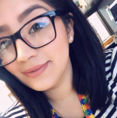 A person wearing glasses and a striped shirt, smiling at the camera. They have dark hair and a colorful lanyard.