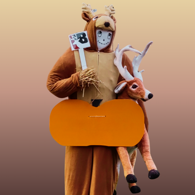 A person in a deer costume holding a deer plush, a sign, and straw, with a large orange shape in front.