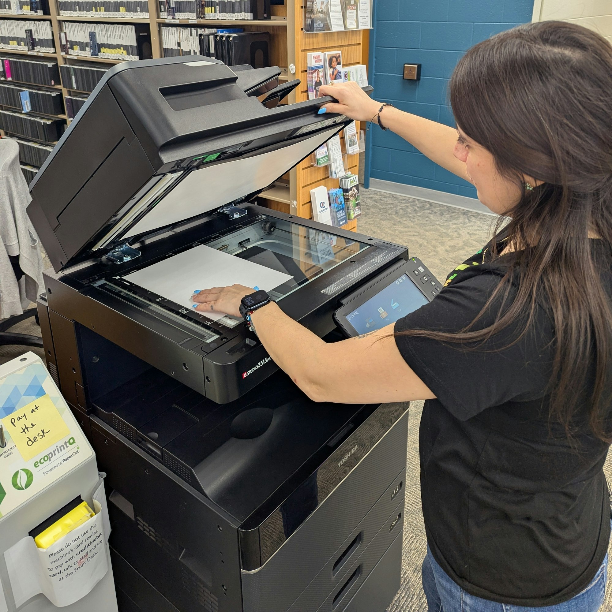A woman is using a printer/scanner in a library, preparing to scan a sheet of paper. Shelves are filled with materials in the background.