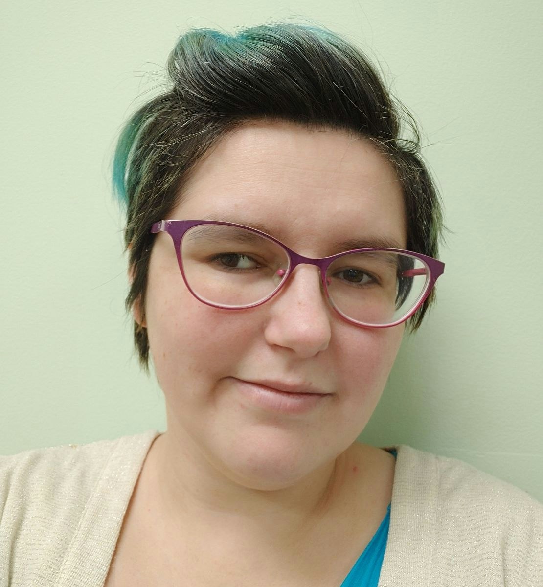 A person with short hair wearing pink glasses and a light sweater, smiling slightly against a light green background.