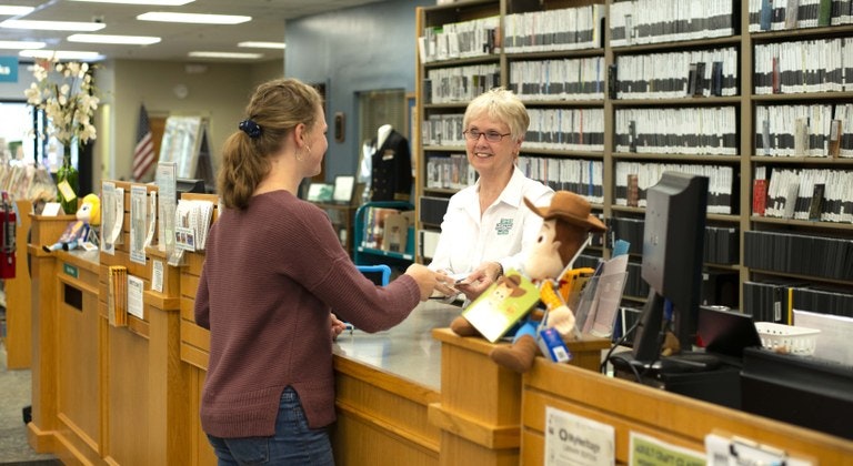 Two people at a library counter, one handing an item to the other. Shelves of books and a computer are visible.