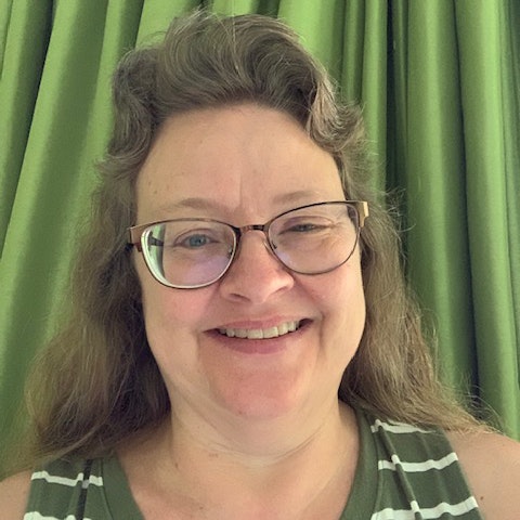 A person smiling, wearing glasses and a striped top, in front of green curtains.