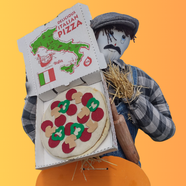 A scarecrow holding a pizza box with an Italian map design, containing a model pizza with felt toppings.