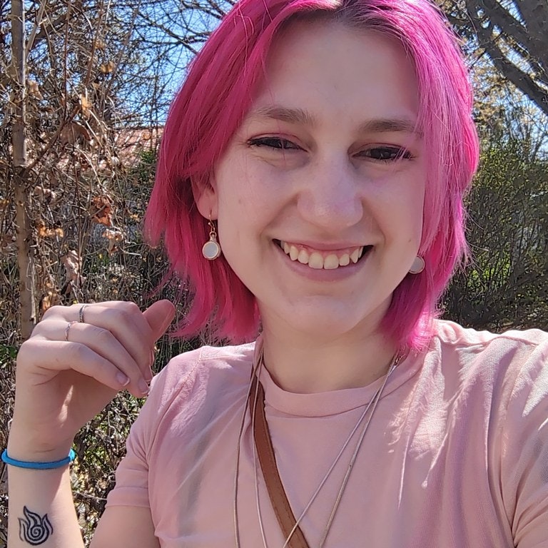 A person with pink hair smiling outside, wearing a pink shirt, necklace, and earrings. A tattoo is visible on their wrist.