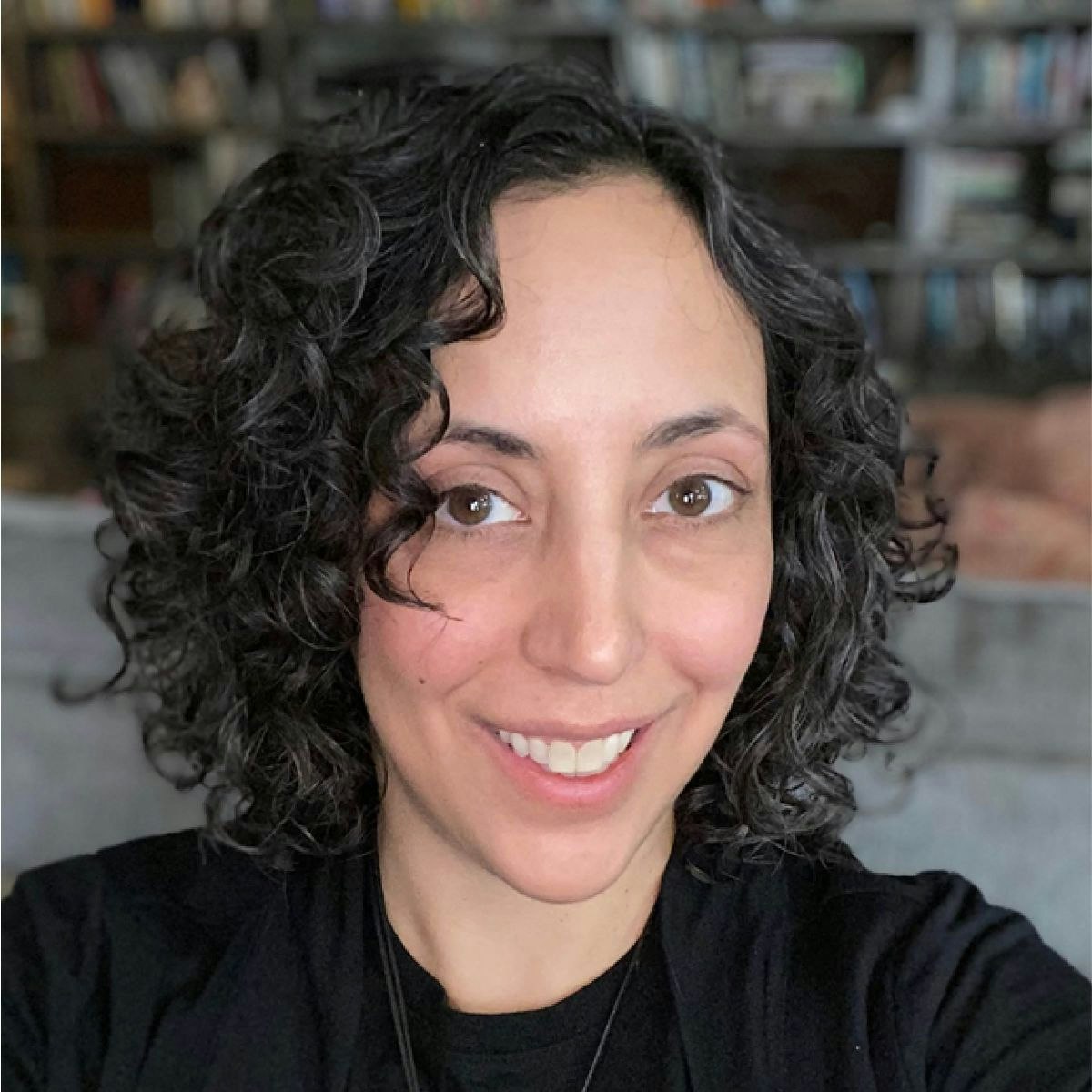 A person with curly hair smiles in a well-lit room, with bookshelves and a sofa in the background.