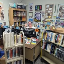 A busy office or library with bookshelves, a desk cluttered with items, and a person waving at the camera.