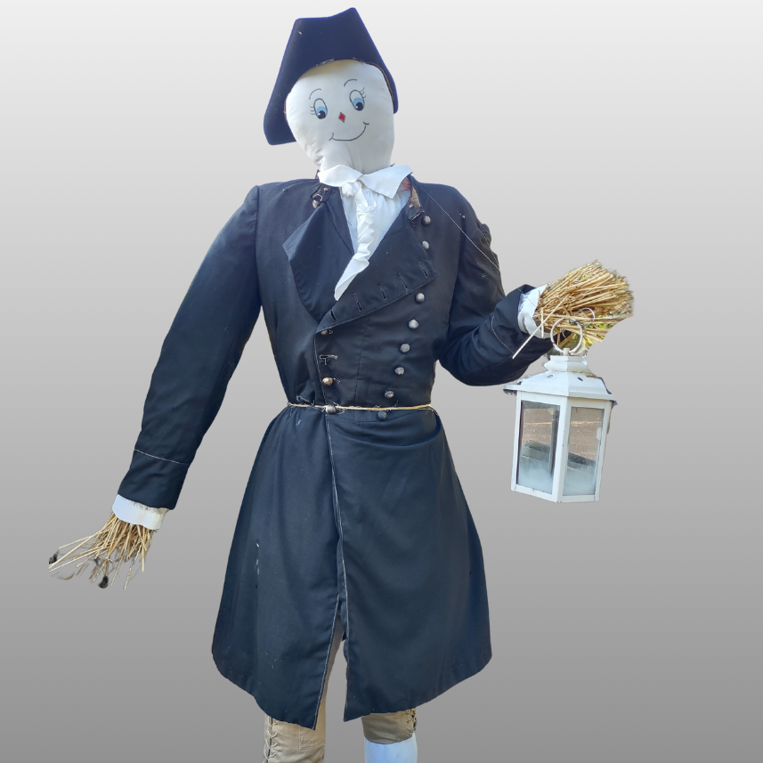 A scarecrow dressed in historic-style clothing, holding a bundle of straw and a white lantern.
