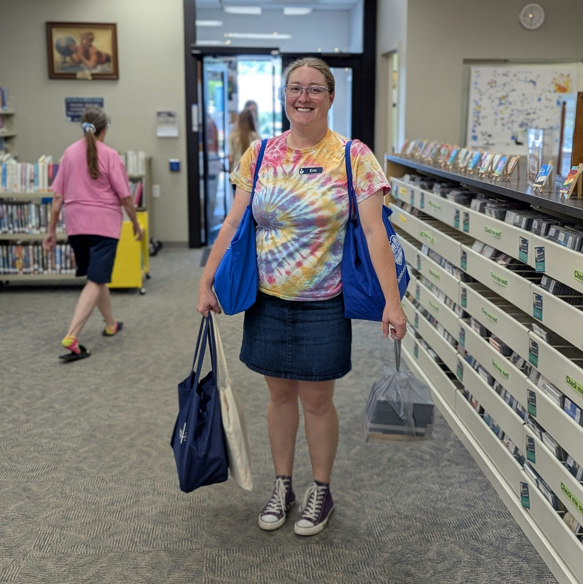 A person in a tie-dye shirt and denim skirt holds bags in a library, with another person walking in the background.